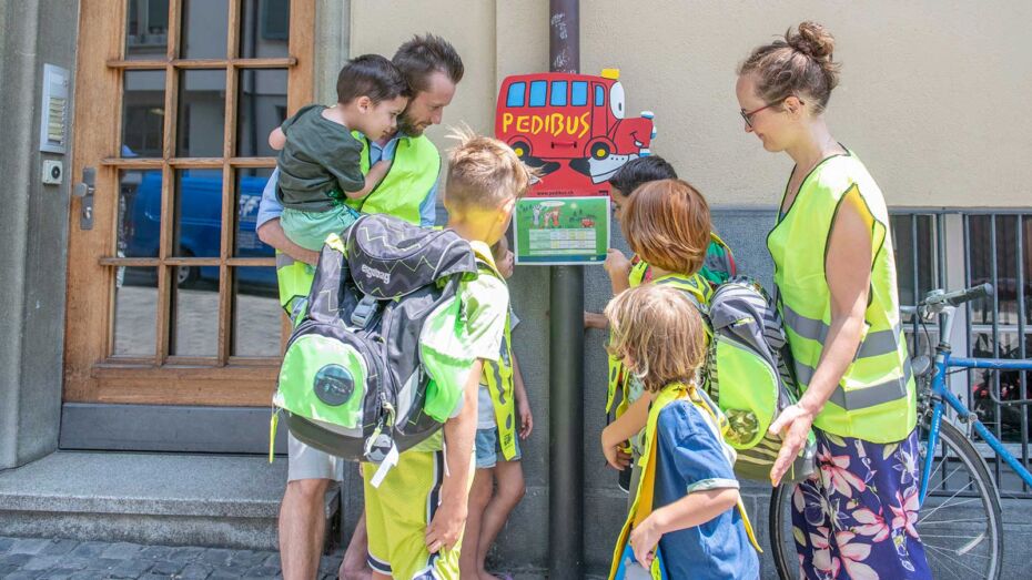 Zwei Erwachsene und sechs Kinder in Leuchtwesten stehen vor einer Pedibus-Haltestelle