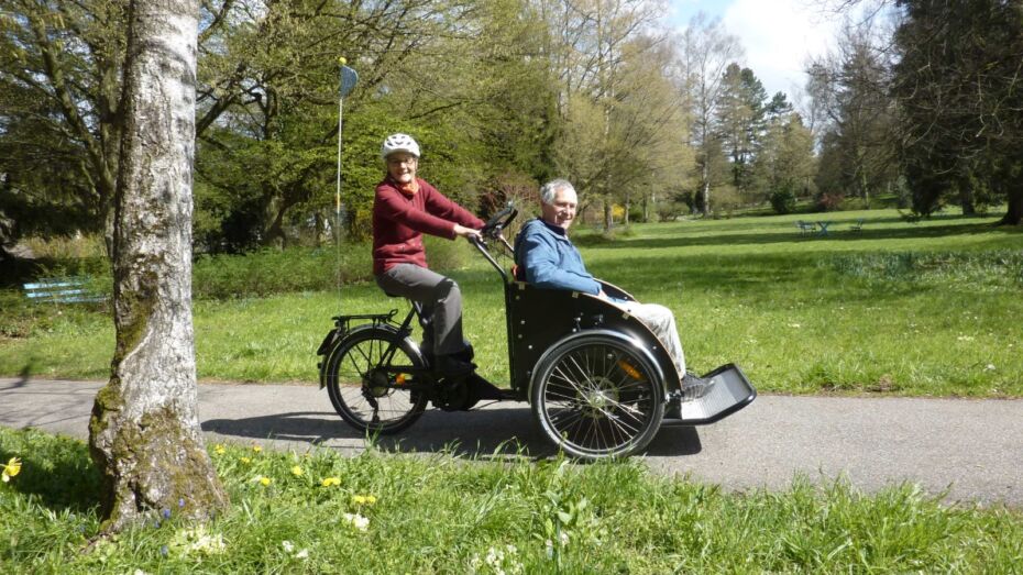 Ein älterer Mann sitzt im Rikscha des Vereins Velores. Das Rikscha wird von einer Frau gefahren.